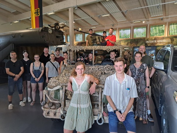 Eine Gruppe von Menschen steht vor einem getarnten Militärfahrzeug in einem Museum. Einige Personen sitzen im Fahrzeug, während andere davor posieren. Im Hintergrund sind weitere militärische Exponate und ein deutsches Flaggenbanner sichtbar.