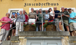 Eine Gruppe von 15 Personen steht auf einer Treppe vor einem Gebäude mit der Aufschrift "Zinzendorfhaus". Die Teilnehmer sind unterschiedlich gekleidet und lächeln in die Kamera. Im Vordergrund stehen Blumenkästen mit Pflanzen.