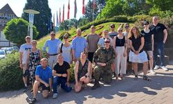 Eine Gruppe von etwa 20 Personen steht im Freien, umgeben von Pflanzen und Fahnen. Einige tragen Freizeitkleidung, während ein Mitglied in Militäruniform ist. Die Gruppe lächelt und posiert für das Foto.