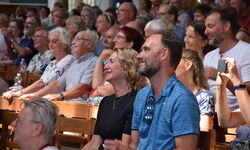 Eine große Menschenmenge sitzt in einem Saal und schaut lächelnd auf die Bühne. Einige Personen klatschen, während andere entspannt wirken. Die Zuschauer sind vielfältig und scheinen an einer Veranstaltung teilzunehmen. (Text KI generiert)